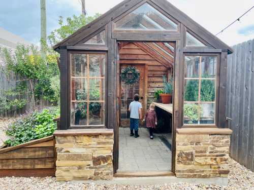 Two children stand inside a greenhouse, surrounded by plants and wooden structures, with a cloudy sky above.