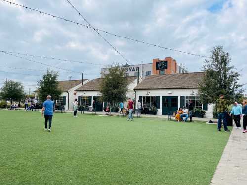 A lively outdoor area with people walking on green grass, surrounded by buildings and string lights under a cloudy sky.