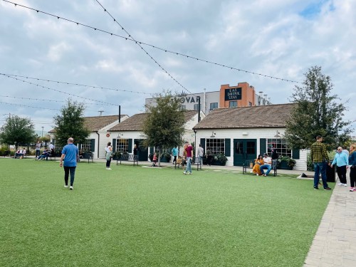 A lively outdoor area with people walking on green grass, surrounded by buildings and string lights under a cloudy sky.