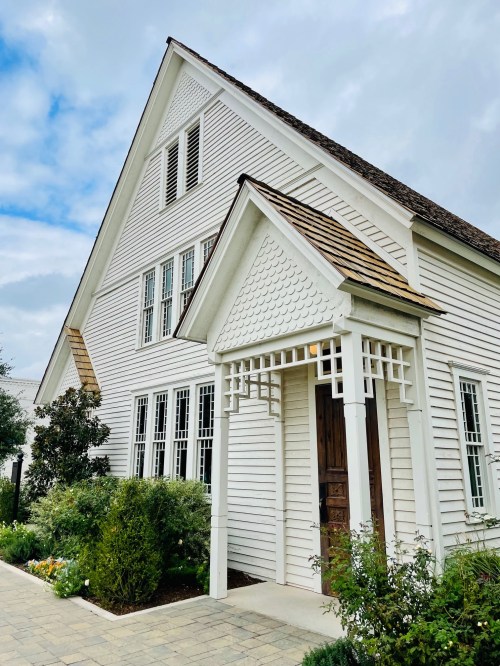A charming white house with a steep roof, large windows, and a welcoming entrance surrounded by greenery.