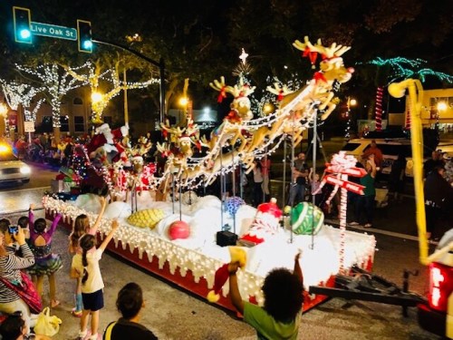 A festive parade float decorated with reindeer, ornaments, and lights, with excited spectators waving nearby.