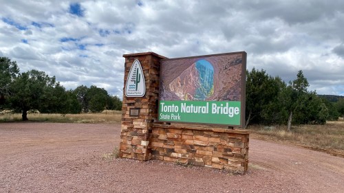Sign for Tonto Natural Bridge State Park, featuring a colorful image of the bridge and surrounded by trees and clouds.