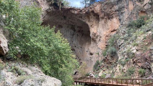 A rocky canyon with a large overhang and lush greenery, featuring a wooden walkway in the foreground.