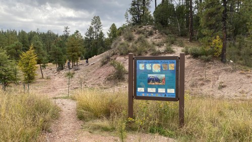 A trail leads to an informational sign about Indian Gardens Paleo Site, surrounded by trees and grassy terrain.
