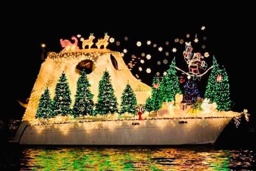 A brightly lit boat decorated with Christmas lights, featuring trees, reindeer, and festive ornaments on a dark water backdrop.