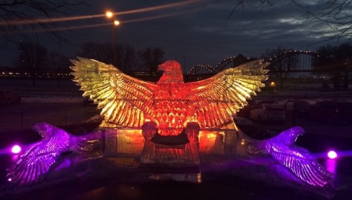 A large illuminated ice sculpture of an eagle with outstretched wings, set against a dark, cloudy sky.