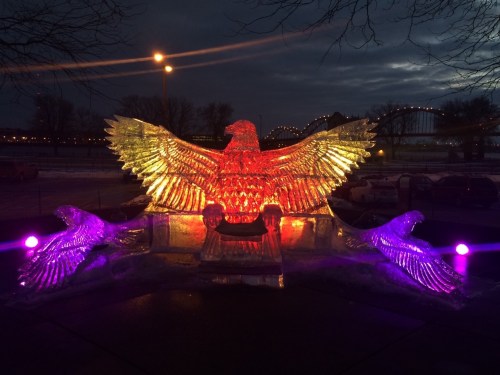 An illuminated ice sculpture of an eagle with outstretched wings, surrounded by colorful lights at dusk.
