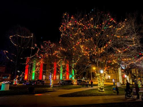 Colorful holiday lights adorn trees and buildings in a festive park at night, creating a cheerful atmosphere.
