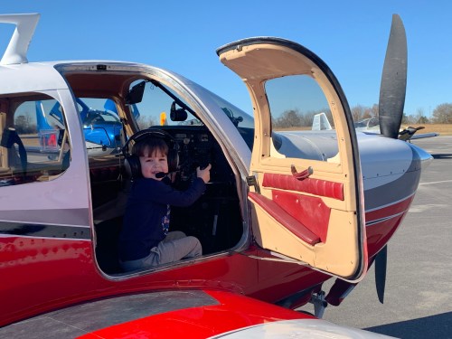 A young child sits in the cockpit of a small airplane, wearing headphones and smiling, with the door open.