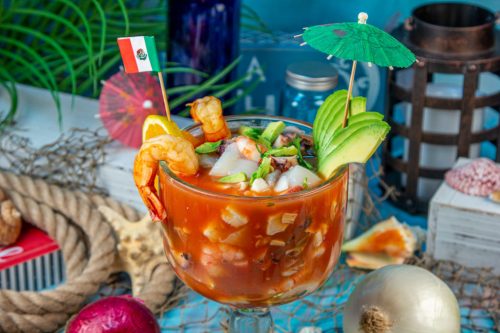 A vibrant seafood cocktail in a large glass, garnished with shrimp, avocado, and decorative umbrellas, set against a colorful background.
