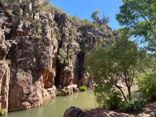 A serene river flows between rocky cliffs, surrounded by greenery under a clear blue sky.
