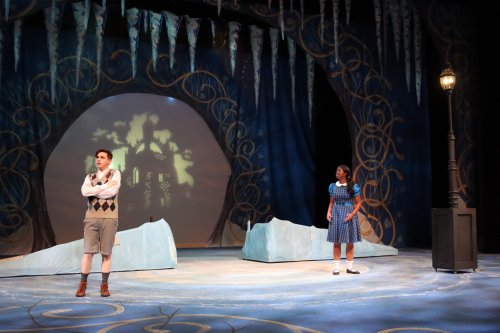 Two actors on stage in a theatrical setting, with icy props and a whimsical backdrop. One looks contemplative, the other attentive.