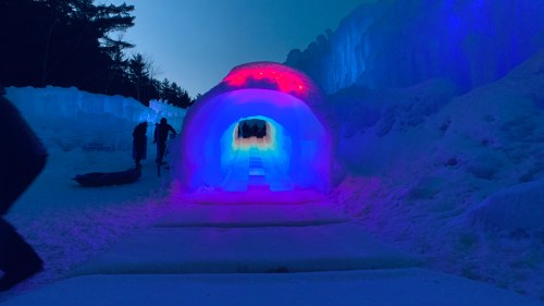A colorful ice tunnel illuminated with blue and red lights, surrounded by snow and trees in a winter landscape.