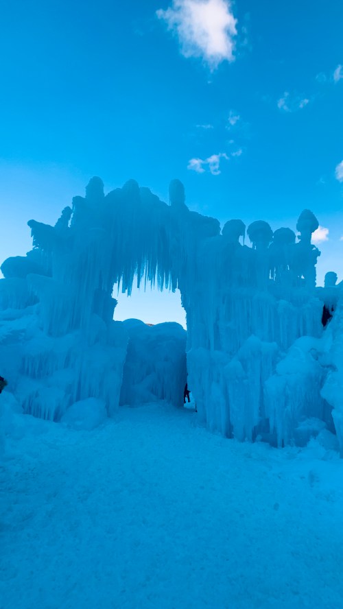 A towering ice arch with icicles against a bright blue sky, with a person walking through the icy passage.