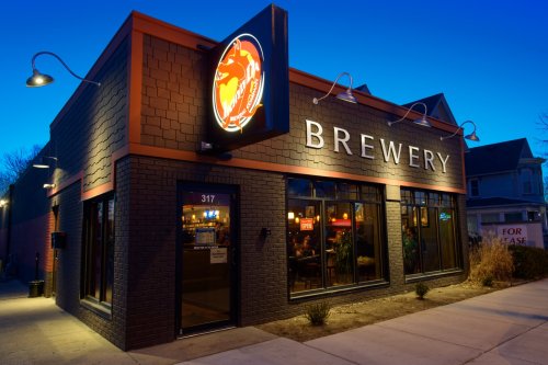 A brewery building at dusk, featuring large windows and a glowing sign, with a sidewalk in front.