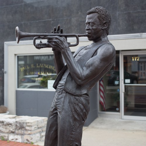 A bronze statue of a musician playing the trumpet, set against a gray building backdrop.