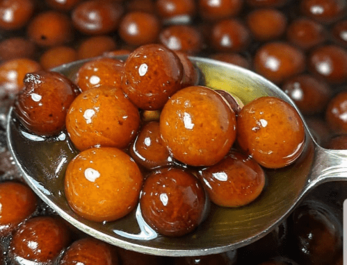 A spoonful of round, glossy brown sweets soaked in syrup, surrounded by more sweets in a dark liquid.
