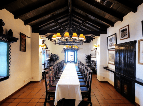 A spacious dining room with a long table, wooden beams, and decorative lighting, featuring a bright window at the end.