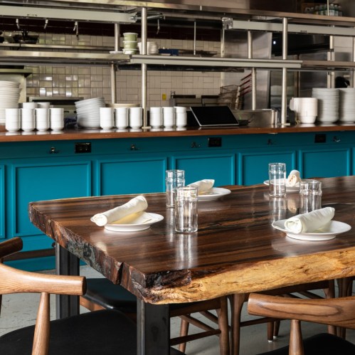 A wooden dining table with glasses and napkins, set in a modern restaurant with a turquoise counter in the background.
