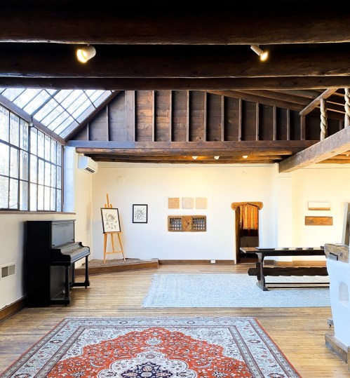 A spacious, well-lit room with wooden beams, a piano, artwork on the walls, and a patterned rug on the floor.