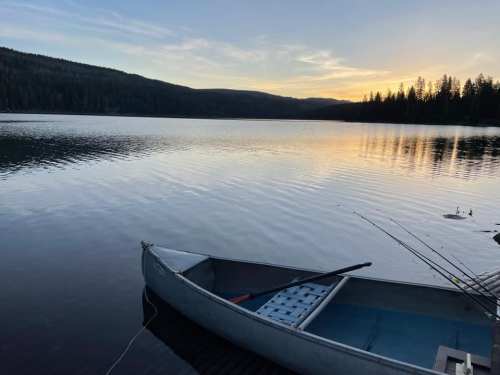 A calm lake at sunset with a canoe moored at the shore, surrounded by trees and mountains in the background.
