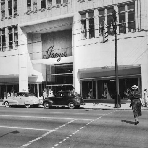 A vintage street scene featuring a department store entrance, classic cars, and a woman walking by in a hat.