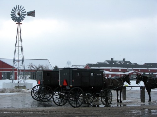 A windmill stands near horse-drawn buggies in a rural setting with red barns in the background under a cloudy sky.