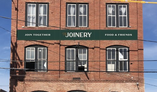 A brick building with a sign reading "The Joinery Food & Friends" and several windows, some with open shutters.