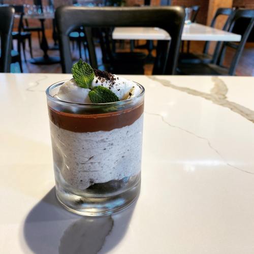 A glass dessert layered with chocolate, cream, and mint leaves, set on a marble table in a restaurant.