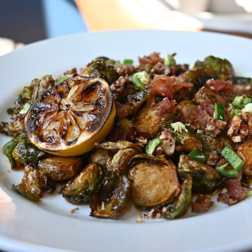 A plate of roasted Brussels sprouts with crispy bits, topped with green onions and a charred lemon slice.