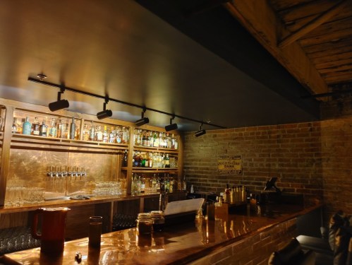 A dimly lit bar with a wooden counter, shelves filled with bottles, and a brick wall in the background.