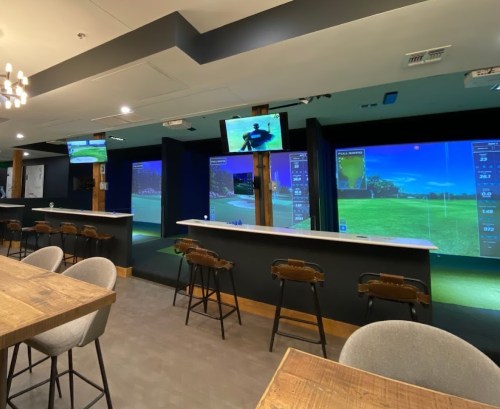 A modern indoor golf facility with multiple screens displaying golf simulations and a bar area with seating.