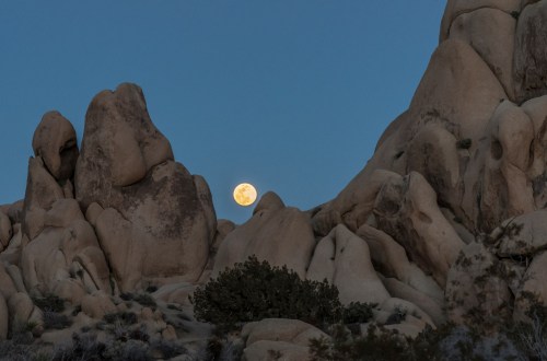A full moon rises above rugged rock formations in a twilight sky.