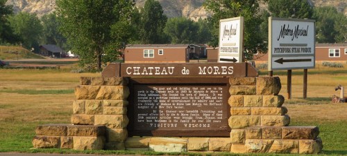 Sign for Chateau de Mores, a historic site, with surrounding greenery and buildings in the background. Visitors welcome.