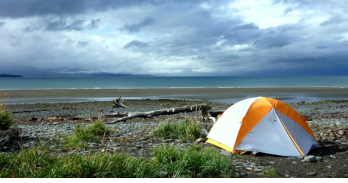 A bright orange tent on a rocky beach with a cloudy sky and calm ocean in the background.