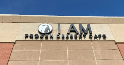 Sign for "I AM Frozen Dessert Cafe" with a penguin logo against a blue sky.