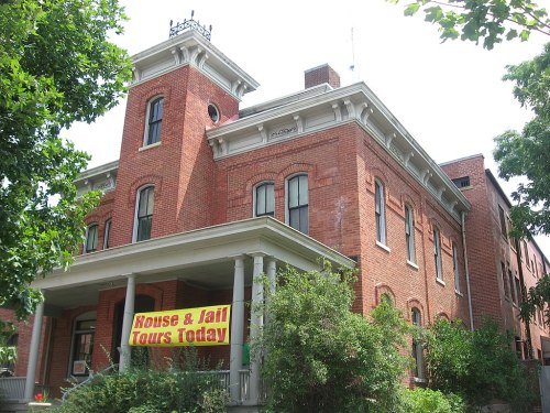 Historic brick building with a tower, featuring a sign for "House & Jail Tours Today" and surrounded by greenery.