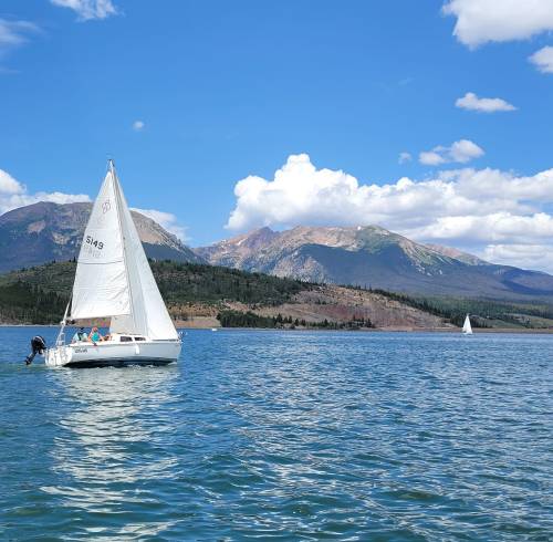 A sailboat glides across a lake with mountains and blue skies in the background.