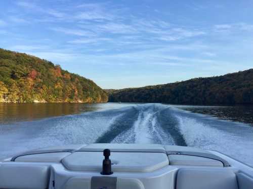 A boat's wake trails behind on a calm lake, surrounded by colorful autumn foliage and rolling hills.