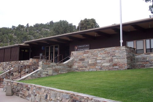 A stone and wood visitor center building with steps, surrounded by greenery and hills in the background.