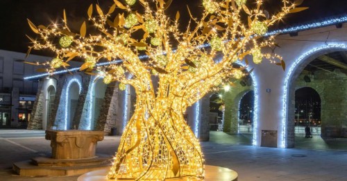 A beautifully lit golden tree sculpture adorned with lights, set in a plaza with arches and festive decorations.