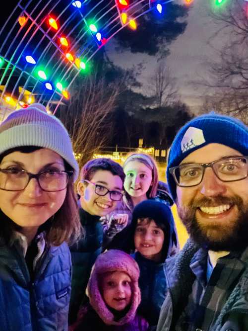 A smiling family poses together under colorful holiday lights at night.