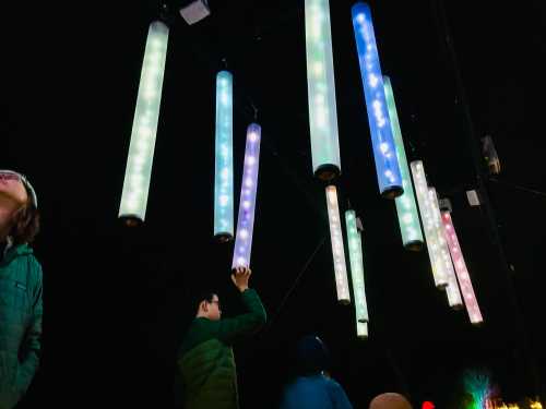 People admire colorful, illuminated vertical tubes hanging from a structure against a dark background.