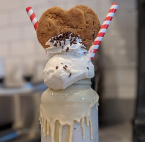 A decadent milkshake topped with whipped cream, a heart-shaped cookie, and two striped straws, with drips of cream.