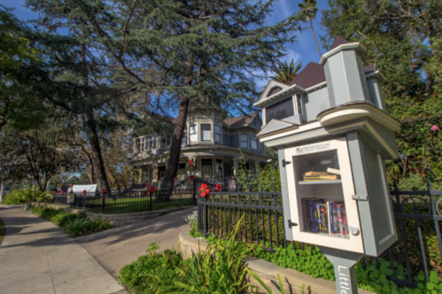 A charming Victorian house with a little free library in the foreground, surrounded by greenery and trees.