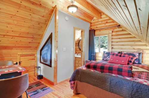 Cozy wooden cabin interior with a bed, small dining area, and a window, featuring rustic decor and warm lighting.