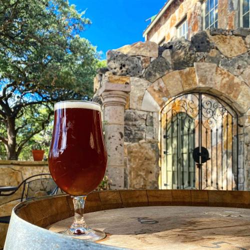 A glass of amber beer sits on a wooden barrel in front of a stone building and lush greenery under a blue sky.
