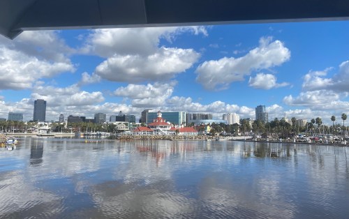 A scenic view of a harbor with reflections, featuring clouds, buildings, and palm trees under a blue sky.