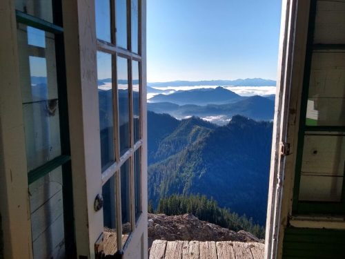 A scenic view of mountains and valleys from an open door, with fog covering the lower landscape under a clear blue sky.