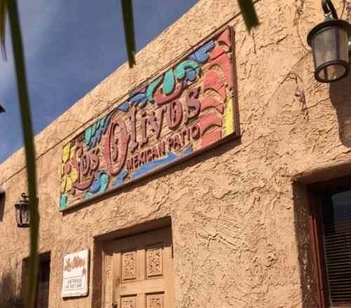 Colorful sign for "Los Olitos Mexican Patio" on a textured beige wall, with decorative elements and outdoor lighting.